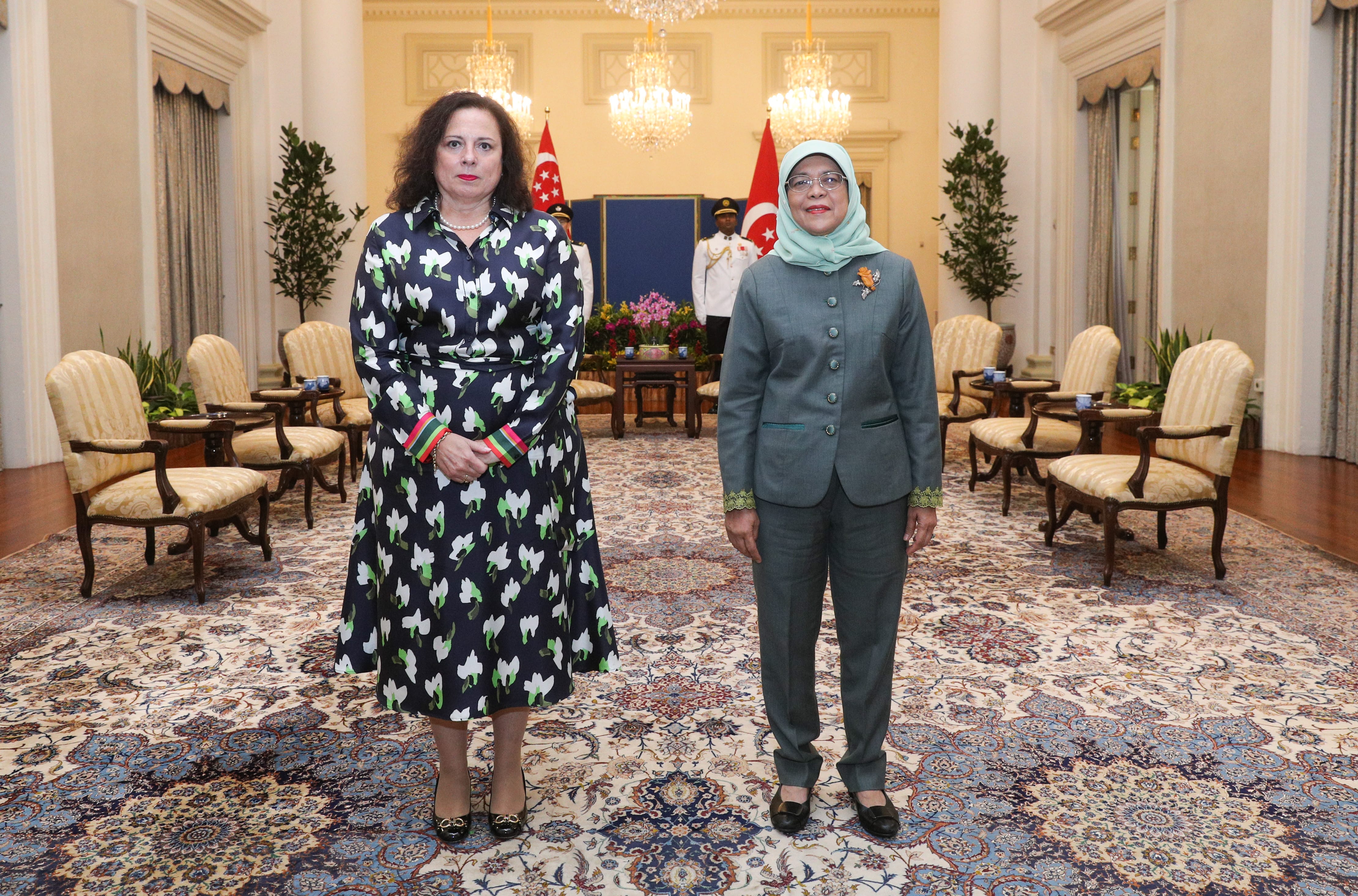 Two women stand indoors on patterned carpet, one in a floral dress, the other in a suit and hijab.